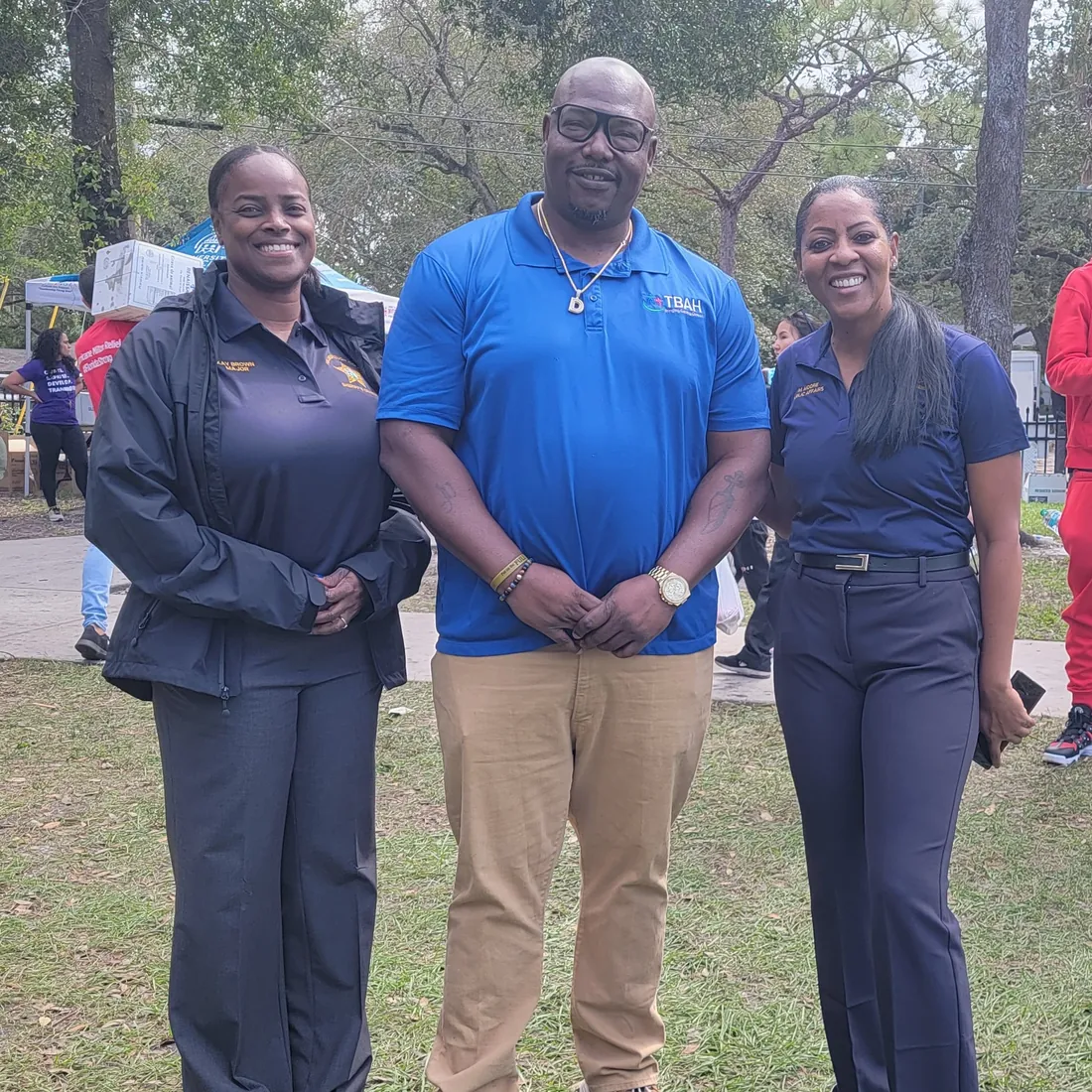 A TBAH staff member in a TBAH polo shirt stands between two uniformed officers at an outdoor community event